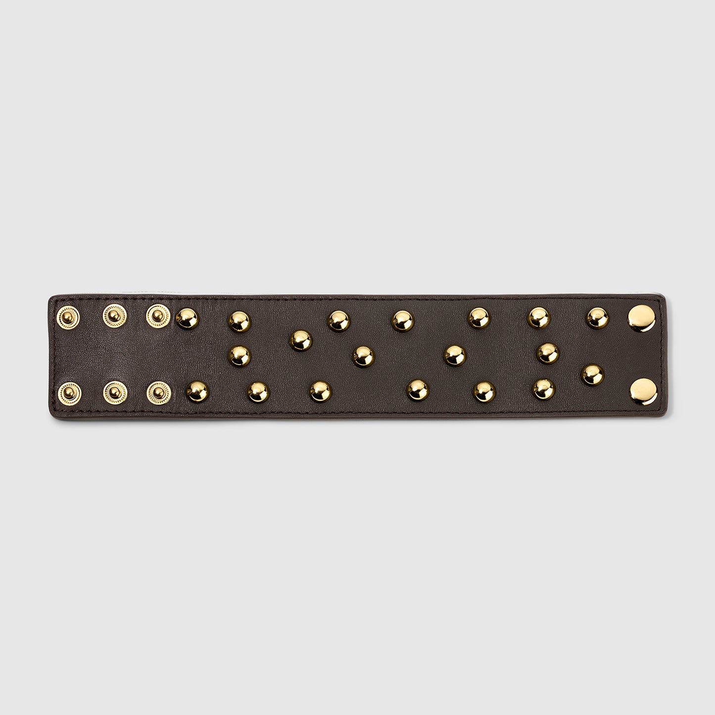 Unfolded view of a brown leather cuff bracelet featuring gold dome studs arranged in rows, with snap-button closures on one side, displayed flat on a light gray background.